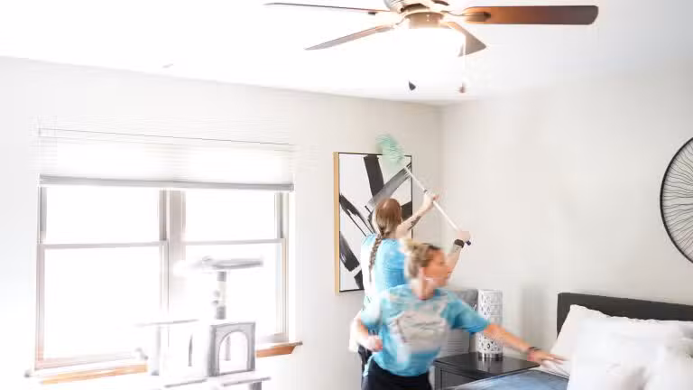 Two cleaners dusting a ceiling fan and wall art in a bright, organized bedroom, illustrating professional maid services in Tulsa for busy families.